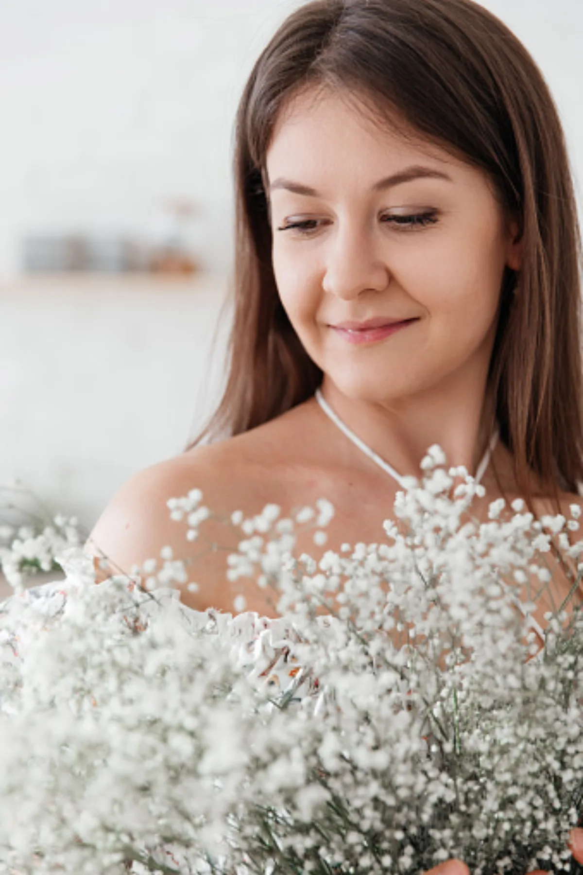 Woman with long hair holding a bouquet of delicate white flowers, smiling in a softly lit interior.