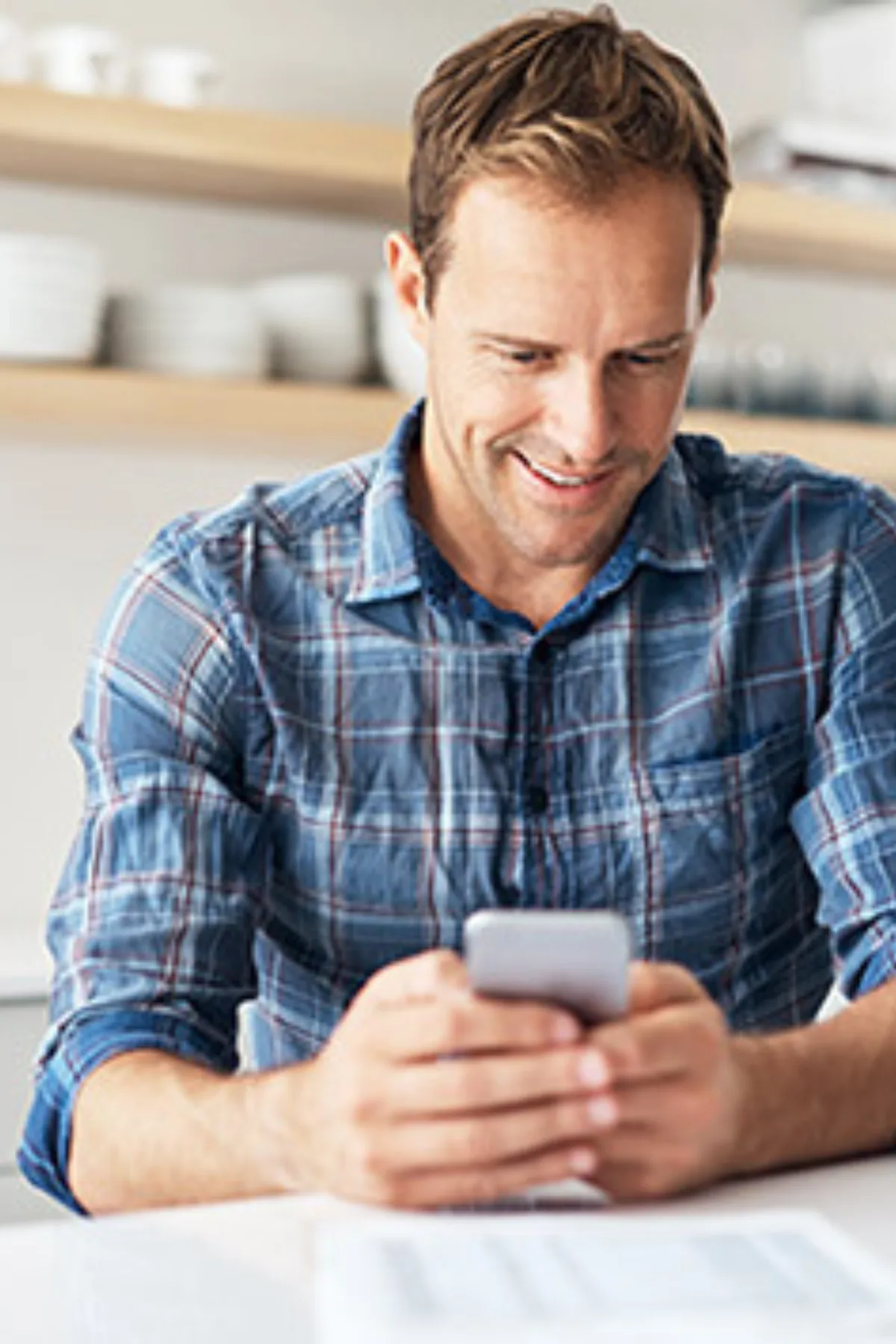 Smiling man in a blue plaid shirt using a smartphone while seated at a table with documents in a bright room.